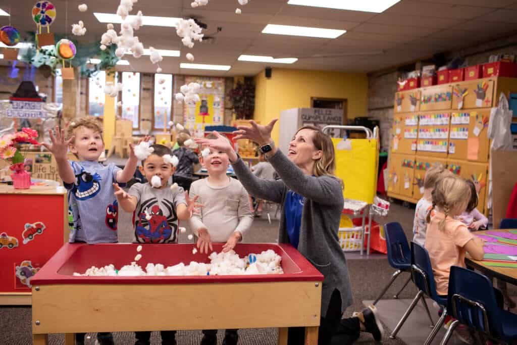 three male preschool students and female teacher throwing cotton balls into air from sensory bin